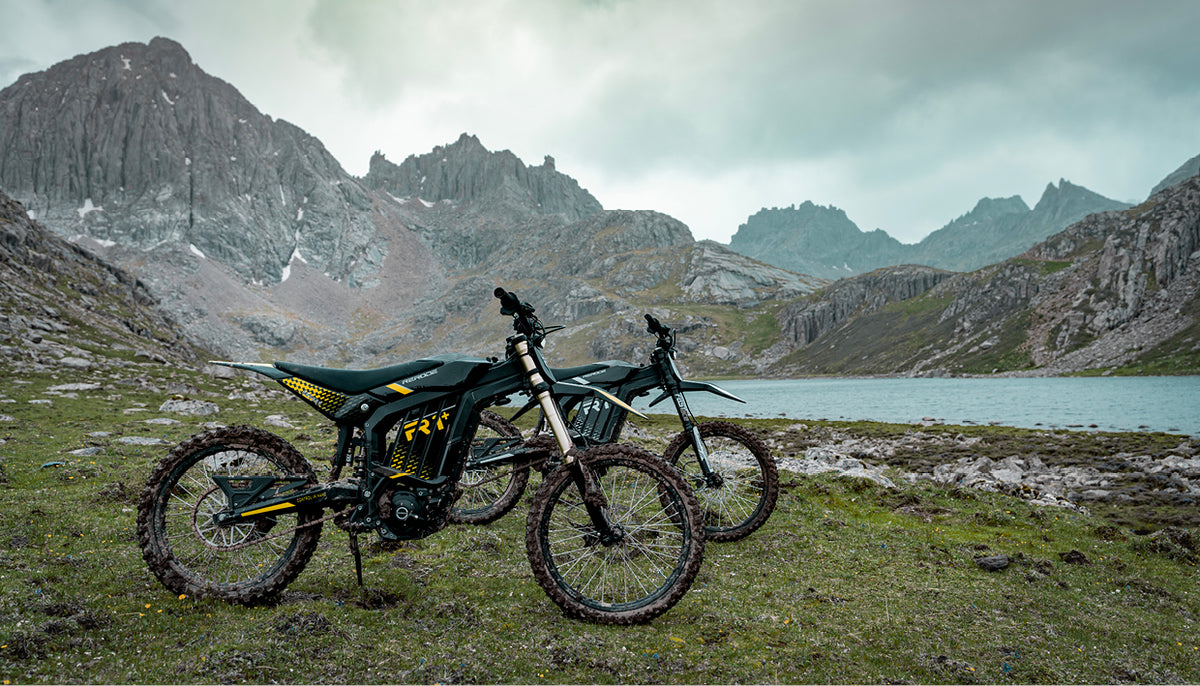 Two Rerode electric dirt bikes beside a mountain lake
